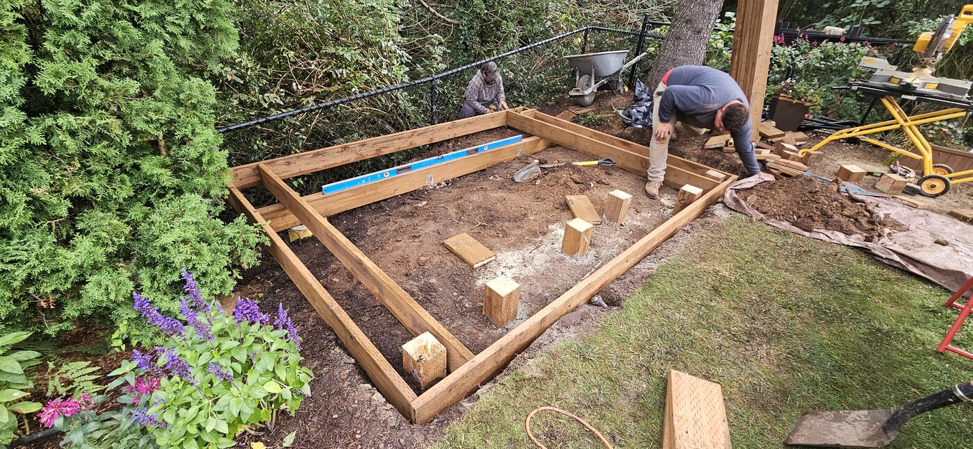 Two crew members squaring a ground-level deck frame beside a flowerbed